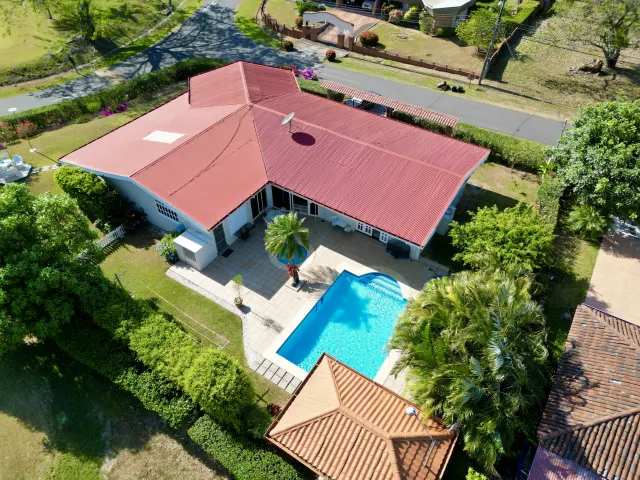 Una vista aérea muestra una casa de una planta con techo metálico rojizo, una piscina en el patio trasero y un patio de losetas rodeado de césped verde.