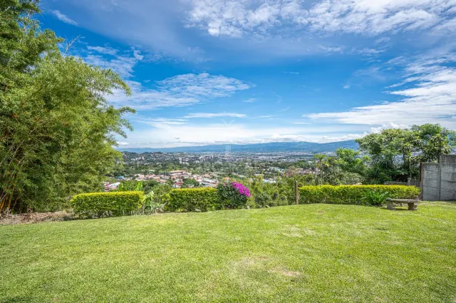Un césped verde abierto con arbustos recortados enmarca una vista panorámica de un valle urbano lejano y montañas bajo un cielo azul con nubes dispersas.