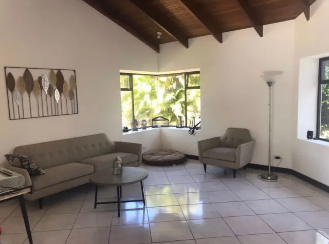 Una sala con paredes blancas, pisos de baldosas, un sofá y un sillón grises, una mesa de centro redonda y ventanas con vista a vegetación exterior.