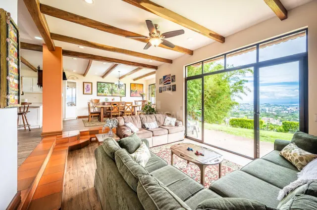 Sala con sofás y mesa de centro junto a grandes puertas de vidrio que muestran un paisaje verde. Unos escalones conducen a un comedor con mesa y sillas de madera.