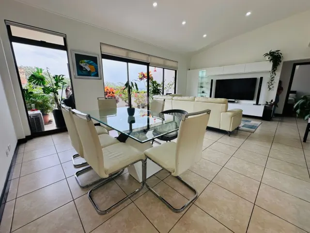 Área de sala y comedor con una mesa de vidrio y sillas blancas, pisos de baldosas, un sofá de cuero blanco y una unidad de televisión en la pared. Amplios ventanales se abren hacia un balcón con plantas.