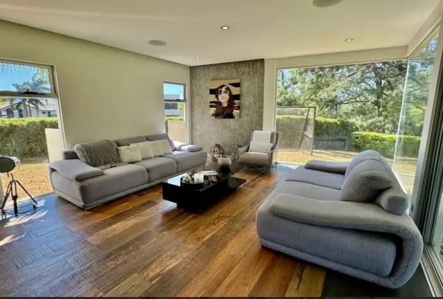 Sala de estar con dos sofás de tela gris, una mesa de centro negra, pisos de tablones de madera y grandes ventanas con vista a la vegetación.