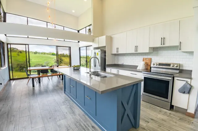 Cocina que cuenta con una isla central azul, electrodomésticos de acero inoxidable, gabinetes blancos y grandes ventanales con vista a colinas verdes.