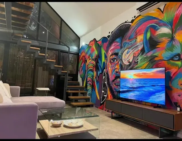 Una sala que presenta un gran mural colorido en la pared, un televisor sobre un mueble multimedia, un sofá y una escalera metálica abierta.