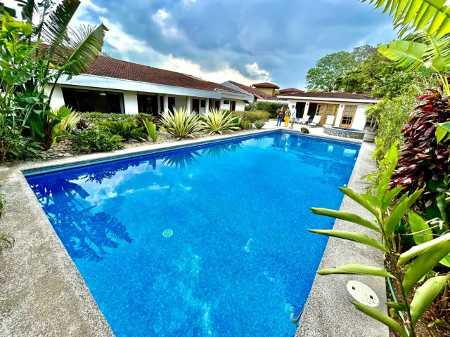 Una piscina rectangular con interior de azulejos azules está rodeada de plantas tropicales y edificios blancos de un piso con techos de tejas bajo un cielo nublado.