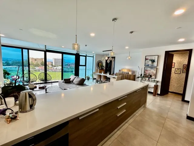 Una larga isla de cocina con gabinetes de chapa de madera y encimera blanca, con vista a un área social abierta con ventanales de piso a techo y vistas al exterior.