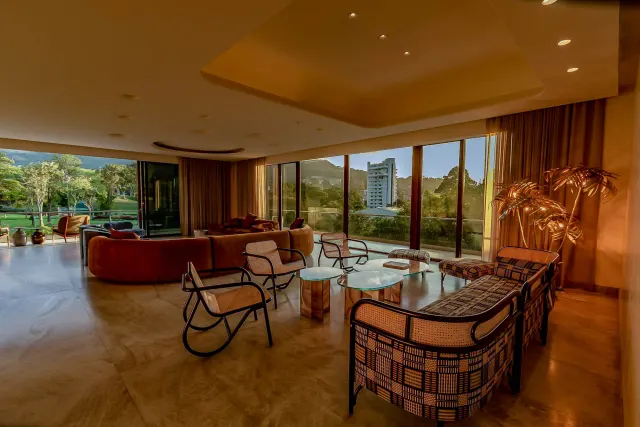 Una amplia sala de estar con un sofá curvo marrón, sillones de ratón, mesas de centro de vidrio y ventanales de piso a techo con vista a la vegetación y edificios.