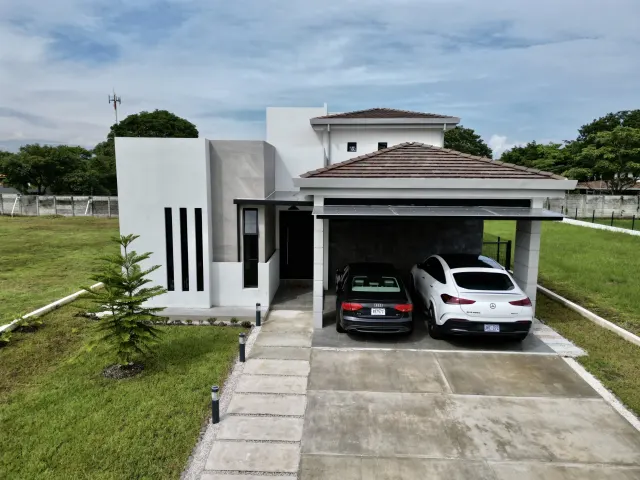 Vista frontal de una casa moderna de dos pisos con paredes exteriores blancas y grises, cochera con techo de tejas y dos autos estacionados.