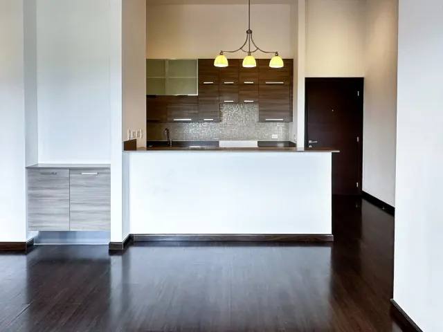 Vista interior de una cocina con gabinetes de madera oscura, una barra desayunadora con base blanca y una lámpara colgante de tres luces.