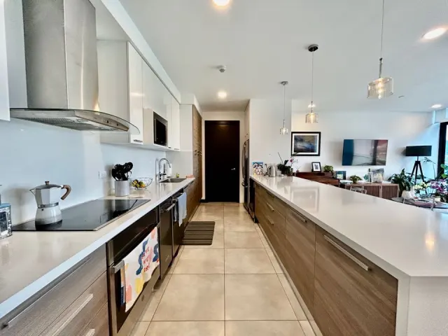 Cocina con una isla larga de encimera de cuarzo blanco, gabinetes de madera, electrodomésticos de acero inoxidable y pisos de baldosas. Al fondo se ve una sala de estar.