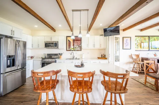 Cocina con una isla central blanca de tres taburetes de madera, gabinetes blancos, electrodomésticos de acero inoxidable y vigas de madera expuestas en el techo.