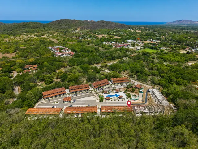 Vista aérea que muestra varios edificios con techos de teja roja, áreas de estacionamiento y una piscina rodeada de árboles verdes y colinas distantes.