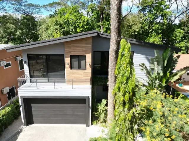Exterior de casa moderna de dos pisos con revestimiento gris, portón de cochera oscuro y balcón en el segundo nivel, rodeada de árboles y vegetación.