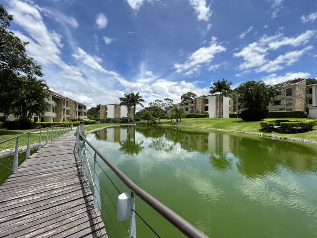 Un puente peatonal de madera con barandillas metálicas cruza sobre un estanque verde, rodeado de edificios de apartamentos y césped bajo un cielo azul.