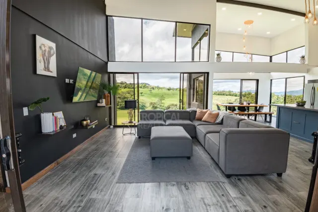 Sala de concepto abierto con un sofá seccional gris, pisos de porcelanato imitación madera gris y cielo raso de doble altura con ventanales que muestran un paisaje verde.
