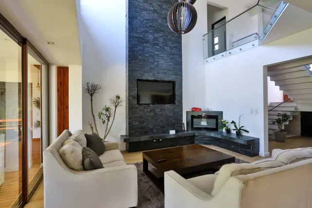 Sala con una pared alta de piedra oscura con televisión integrada, una mesa de centro de madera oscura y dos sofás blancos.