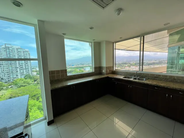 Área de cocina con gabinetes inferiores oscuros, sobres de granito, fregadero de doble cubeta y grandes ventanas con vistas urbanas.