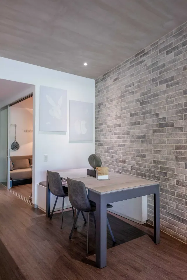 Mesa de comedor y sillas frente a una pared texturizada de ladrillo gris, con vista a una habitación contigua con un sofá.