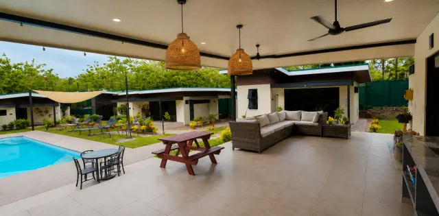 Un patio techado con piso de cerámica cuenta con un sofá seccional de mimbre, una mesa de picnic de madera y lámparas colgantes tejidas, con vista a una piscina y jardín.