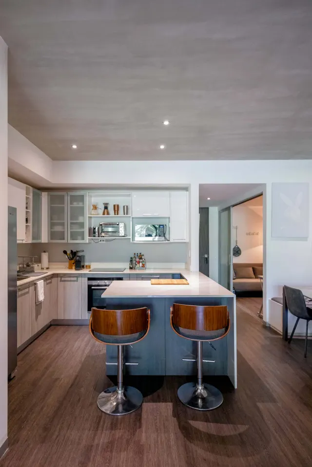 Cocina con pisos de laminado de madera, isla central, dos taburetes de barra, gabinetes blancos y de vidrio, y electrodomésticos empotrados.