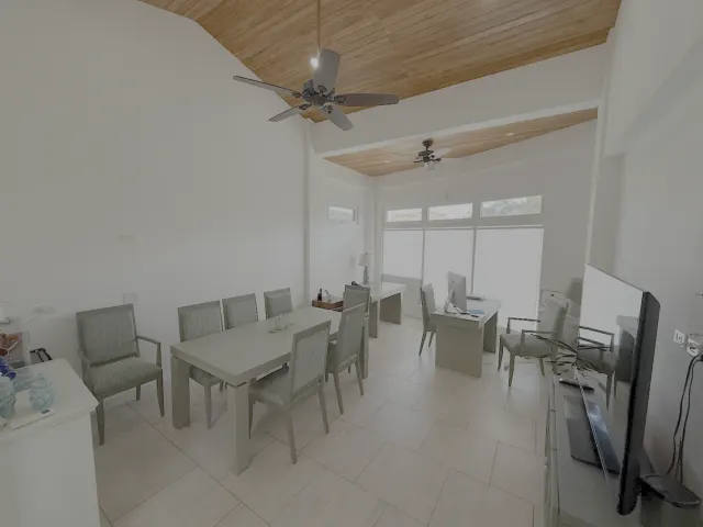 Sala de oficina con una mesa larga y sillas tapizadas, escritorios cerca de ventanales grandes y un cielo raso inclinado de tablones de madera.