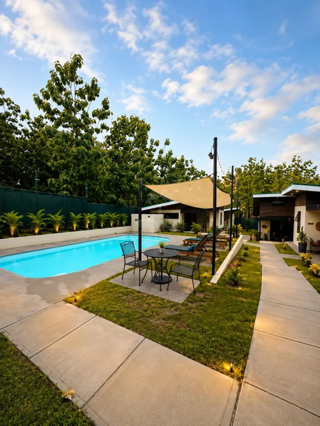 Área exterior con una piscina rectangular, una mesa con cuatro sillas bajo una vela de sombra, tumbonas y pasillos de concreto rodeados de césped e iluminación.