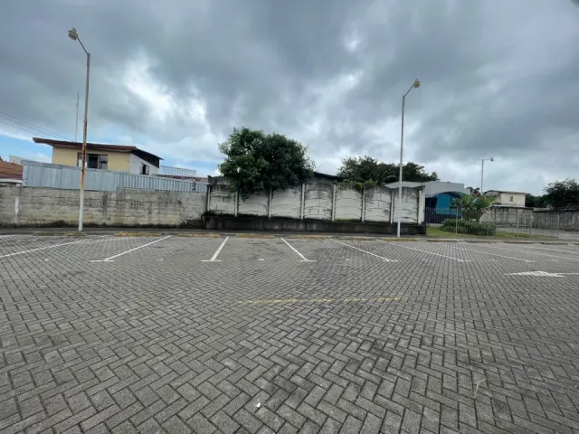 Un estacionamiento al aire libre pavimentado con adoquines grises cuenta con espacios blancos pintados, postes de luz y un muro perimetral bajo un cielo nublado.
