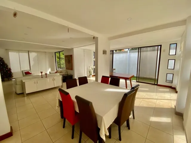 Un comedor de concepto abierto con mesa cuadrada y sillas tapizadas, que conecta con una sala luminosa con sofá blanco y puertas de vidrio.