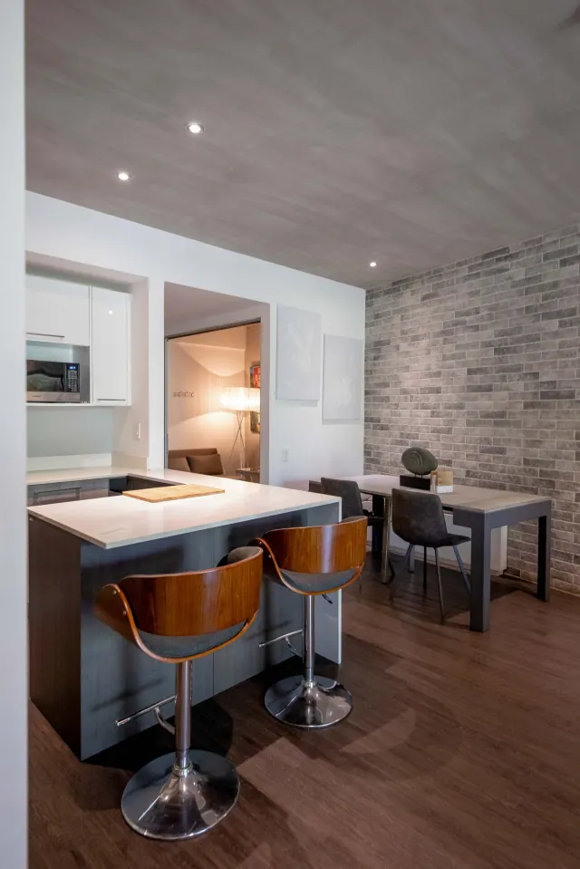 Barra de cocina con dos taburetes de madera frente a una mesa de comedor contigua y una pared de ladrillo gris.