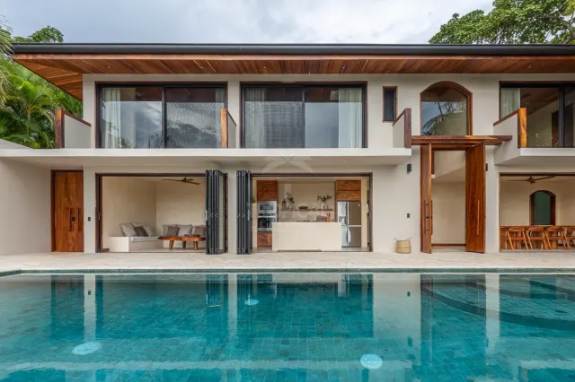 Fachada de casa de dos pisos con una piscina en primer plano, puertas corredizas de vidrio y detalles de madera en el techo.