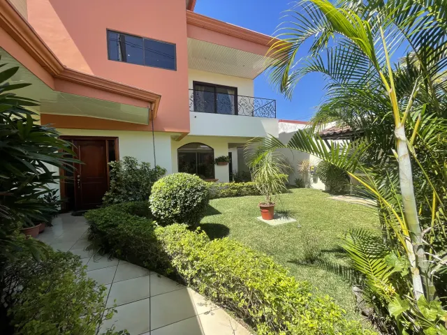 Un césped verde y arbustos ajardinados se encuentran frente a una casa de dos plantas con exterior color durazno y un camino de losetas.