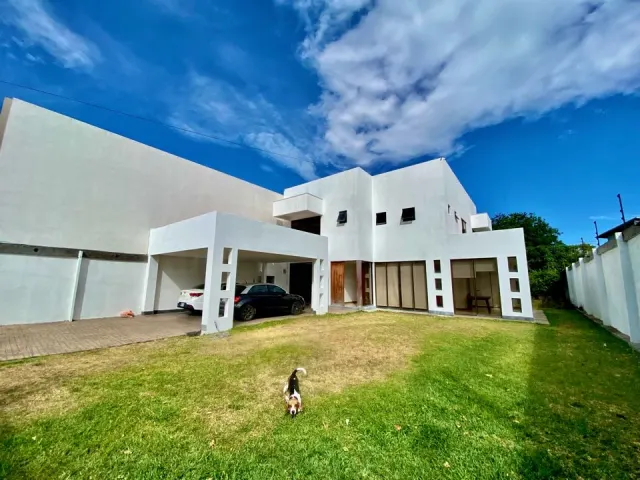Fachada blanca de dos pisos de una casa moderna que incluye una cochera techada con vehículos estacionados y césped al frente bajo un cielo azul.