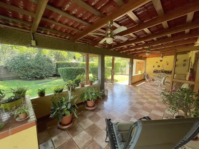 Un patio de baldosas con techo de madera expuesta, ventiladores de techo y pilares de concreto que da a un jardín con césped y plantas en macetas a lo largo de la repisa.