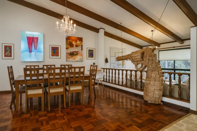 Comedor con mesa de madera y sillas, pisos de parquet, vigas expuestas en el techo y cuadros en la pared. Una gran escultura tejida se encuentra cerca de un barandal de madera.