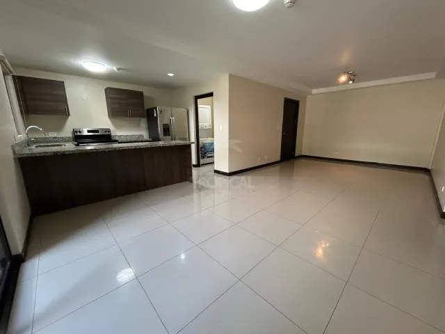 Un espacio interior abierto que incluye una cocina con gabinetes oscuros, sobres de granito y un refrigerador de acero inoxidable. La habitación contigua tiene pisos de cerámica blanca.
