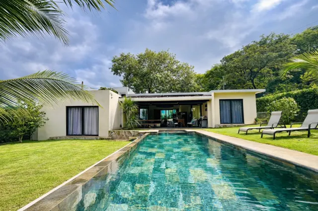 Exterior de una casa de una planta con una piscina de nado alargada en primer plano, rodeada por un césped verde y árboles maduros.