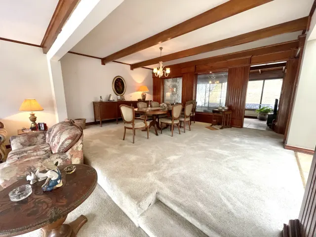 Sala y comedor alfombrados con vigas de madera oscura expuestas en el cielo raso, paneles de madera y una mesa de comedor ovalada con seis sillas.