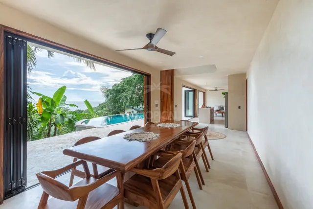 Una terraza abierta y techada que cuenta con una larga mesa de comedor de madera con sillas a juego, ventilador de techo y vista a una piscina exterior y al mar a través de una gran abertura.