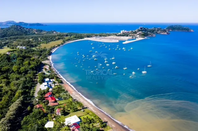 Vista de alta perspectiva de una bahía costera con numerosos botes anclados, una playa de arena y vegetación circundante bajo un cielo despejado.