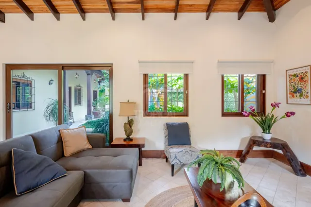 Área de estar con sofá sectional gris, dos ventanas, puerta corrediza de vidrio y una silla decorativa contra paredes blancas.