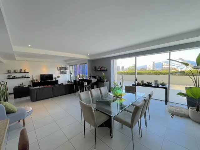 Espacio de sala y comedor con una mesa de comedor de vidrio y seis sillas, un sofá oscuro y amplios ventanales que dan a un balcón con vistas a la ciudad.