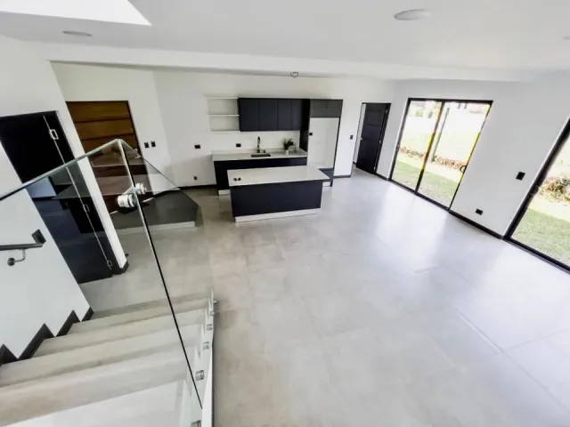 Espacio interior abierto que muestra una cocina con gabinetes oscuros e isla, pisos de baldosas grises y una escalera con barandal de vidrio.
