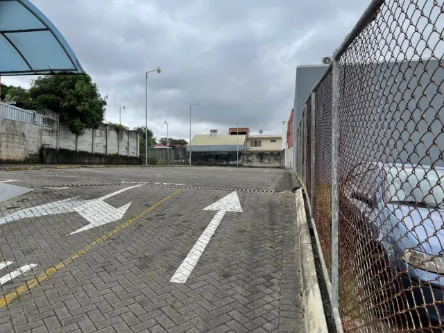 Un estacionamiento pavimentado y vacío con flechas direccionales pintadas en el suelo, cercado con malla metálica y muros de concreto bajo un cielo nublado.