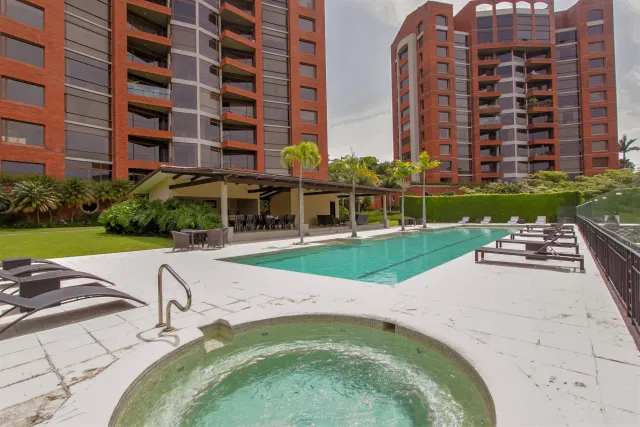 Una piscina al aire libre y un jacuzzi circular en una terraza de baldosas, rodeados de tumbonas, palmeras y edificios de ladrillo rojo.