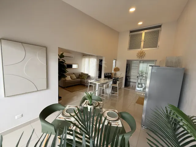 Una vista interior que muestra un comedor en primer plano, una cocina con isla blanca y una sala contigua con sofá.