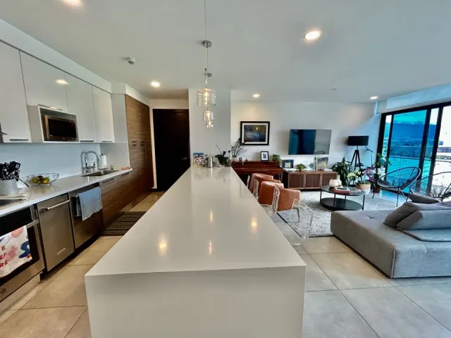 Sala y cocina de concepto abierto con una larga isla de cocina blanca, lámparas colgantes de vidrio y un sofá modular gris.