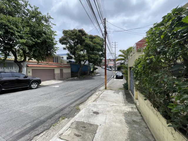 Vista de una calle pavimentada con acera, líneas eléctricas aéreas y vehículos estacionados bajo un cielo nublado.