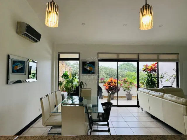 Espacio de sala y comedor con una mesa de comedor de vidrio, sillas blancas, dos lámparas colgantes y puertas corredizas hacia un balcón con plantas.