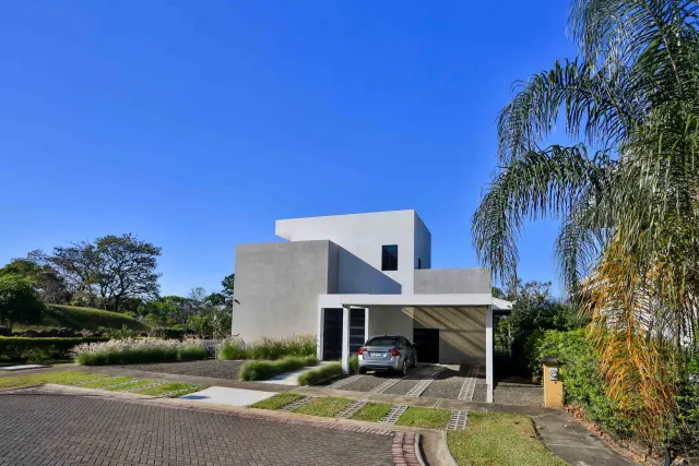 Vista exterior de una casa moderna de dos pisos de concreto con una cochera techada y un auto estacionado, rodeada de césped y árboles bajo un cielo azul despejado.