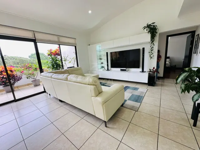 Sala con un sofá de cuero de tonos claros, un mueble de televisión grande montado en la pared y puertas corredizas de vidrio hacia un balcón.